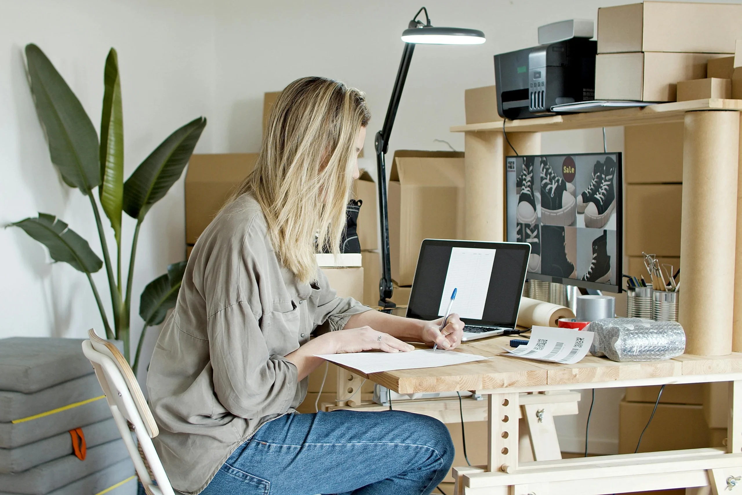 A Woman Writing on Paper with a Tablet placed in Front of The Table. Card boxes Containing Products Placed Throughout the Room. Best Items to Resell for Profit.