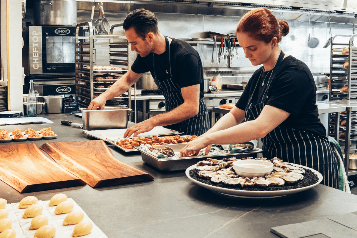 Two individuals arranging food on trays in a commercial kitchen, representing the steps on how to start a catering business.