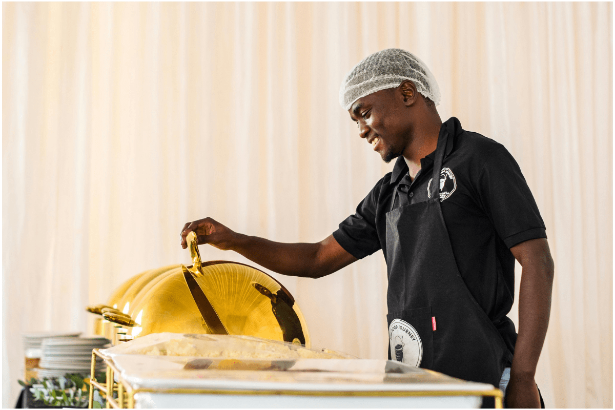 A catering server standing near a serving table, preparing to lift the lid of food dishes at an event, illustrating a key aspect of how to start a catering business