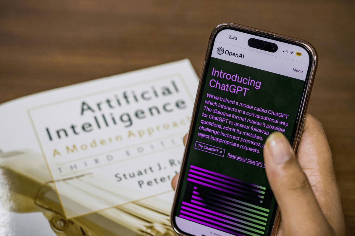 Mobile phone displaying ChatGPT screen held above 'Artificial Intelligence' book on a table – showing how to make money with ChatGPT and AI technology.