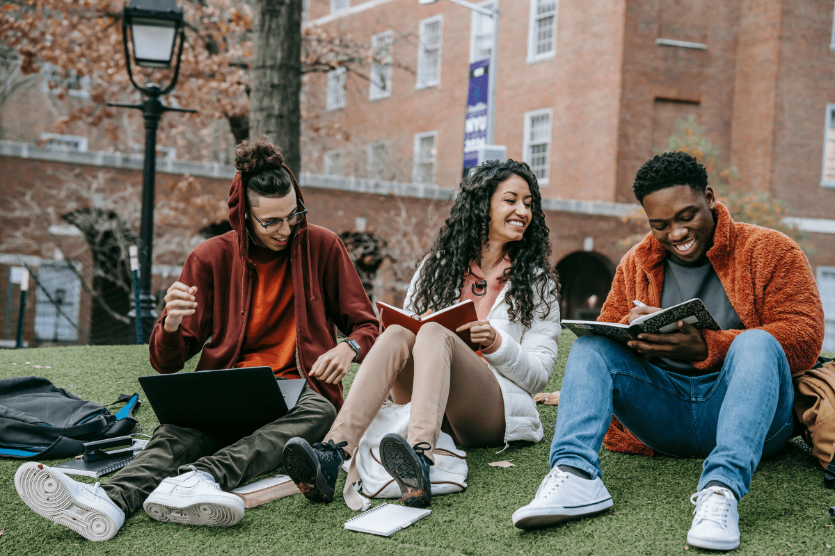 Three college students sitting outdoors, enjoying campus life while working on side hustles. Two students have notebooks open, and one uses a laptop, representing side hustle ideas for college students.