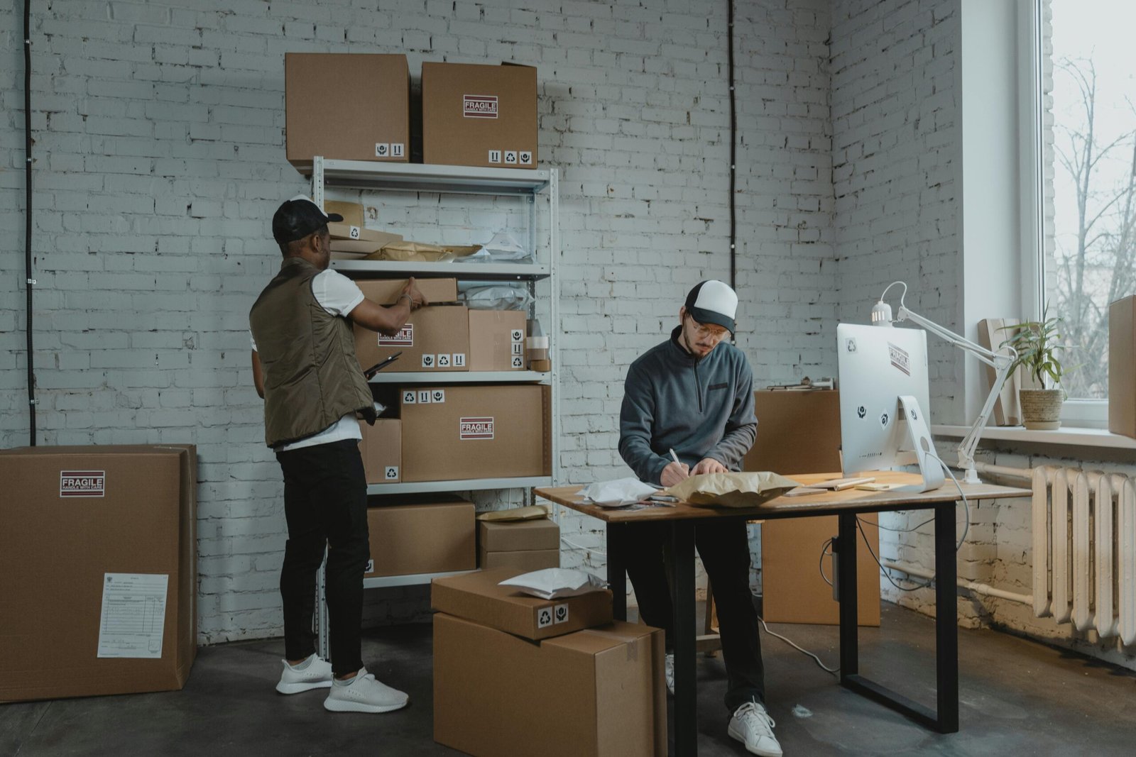 Important Steps on How to Start a Box Truck Business Two persons are in a warehouse where one person is writing notes on how to start a box truck business, while the other lifts a cardboard box from shelves with scattered cardboard boxes around them.