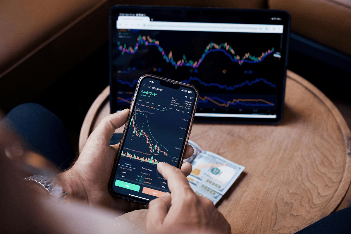 A Person Hand Visible Holding a Phone with a Laptop Sitting on the Table in front with Both Screens Shoing Stocks Going Up and Down Illustrating Best Dividend Stocks for Passive Income