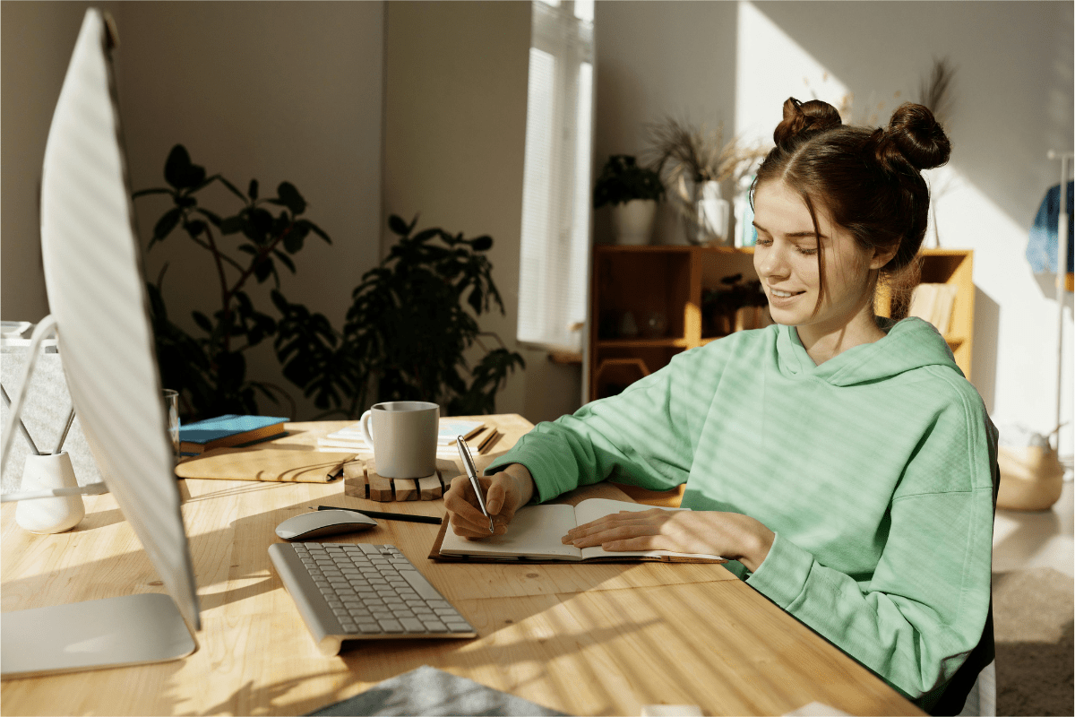 Freelancing - Side Hustles for Teens A Teenage Girl Can be Seen Sitting in front of laptop Writing Something in Her Book Illustrating Side Hustles for Teens