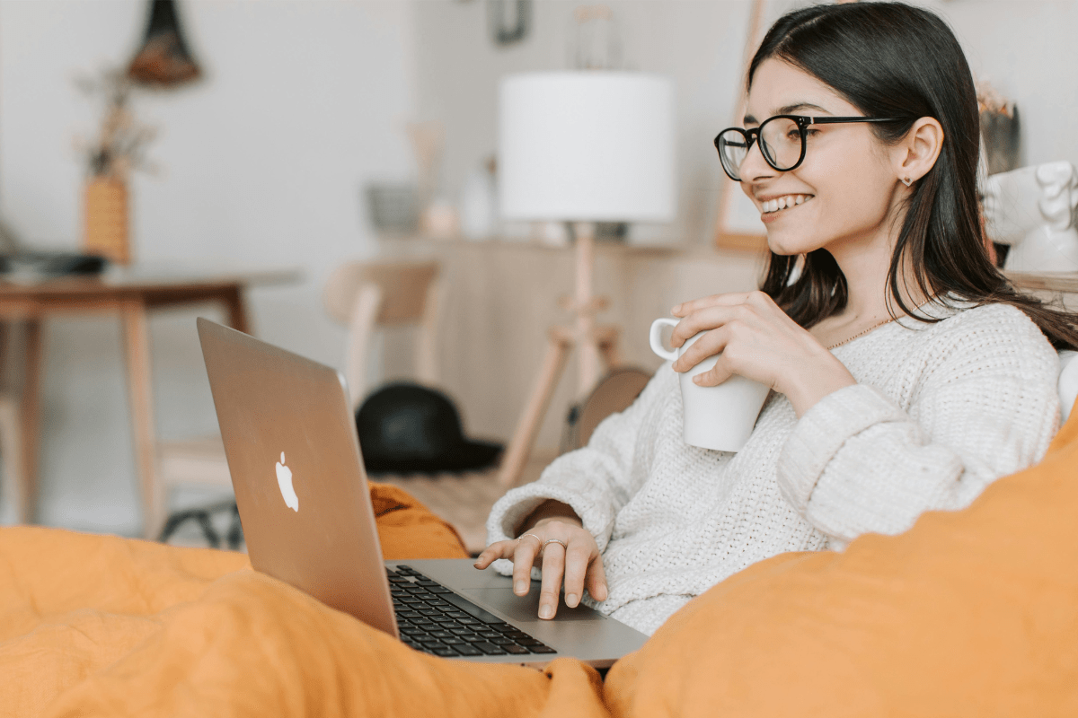 A Woman Sitting on a Couch Holding a Coffee Mug with Her MacBook Placed In Front Illustrating Side Hustles that Pay Daily