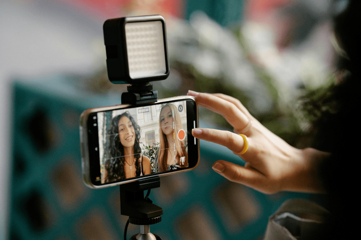 Two Women Can be Seen on The phone Recording Themselves while a Woman is Pressing the Stop Button. The Phone is Set on a Tripod Stand Illustrating How to Make Passive Income On YouTube.