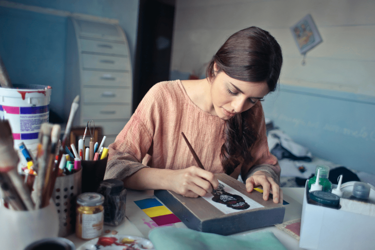 A Woman Can Be seen Painting on a Canvas Placed on Table Illustrating Side Hustles for single moms.