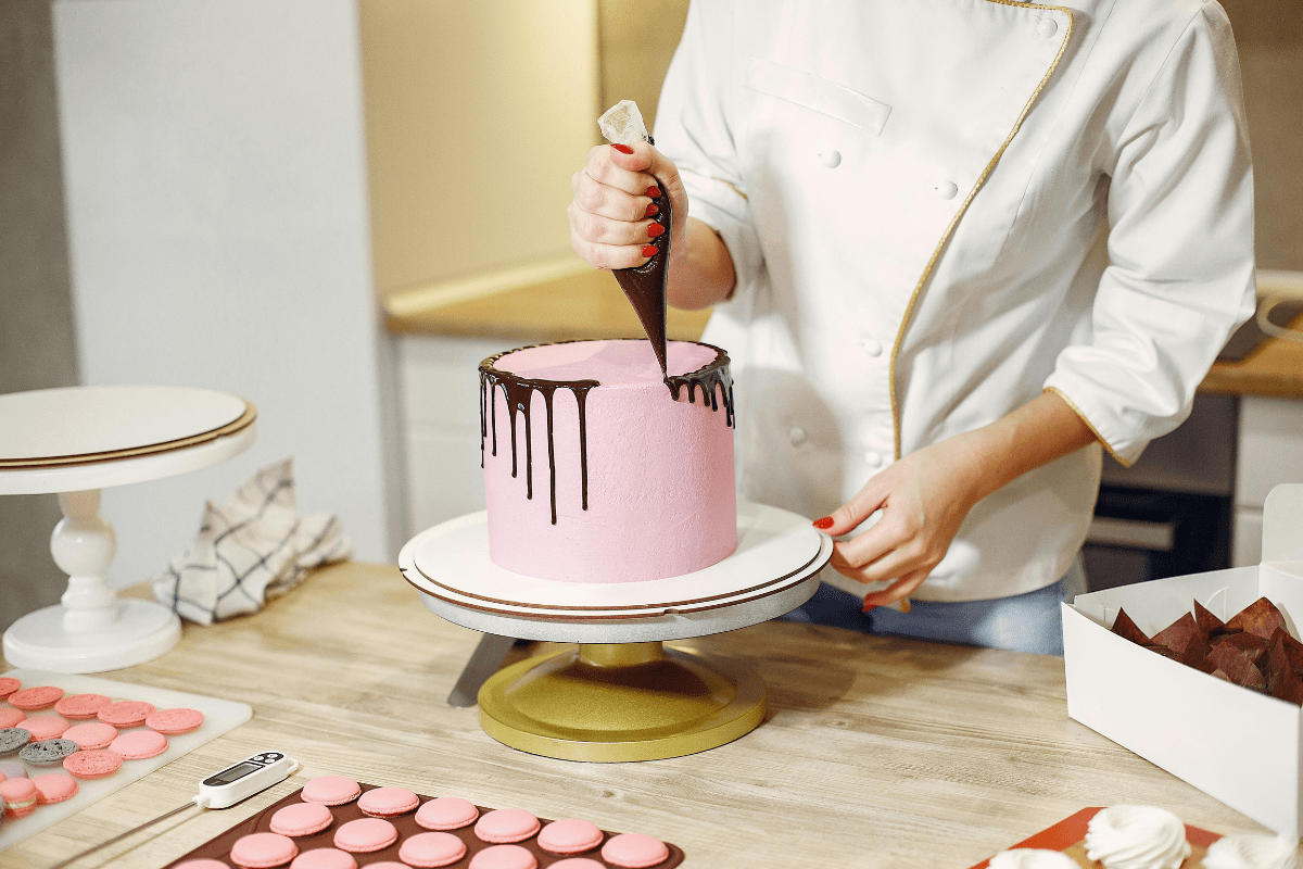 A Woman's Hands Are Visible Holding a Piping Bag Decorating the Cake Placed In Front Illustrating Side Hustles for Single Moms