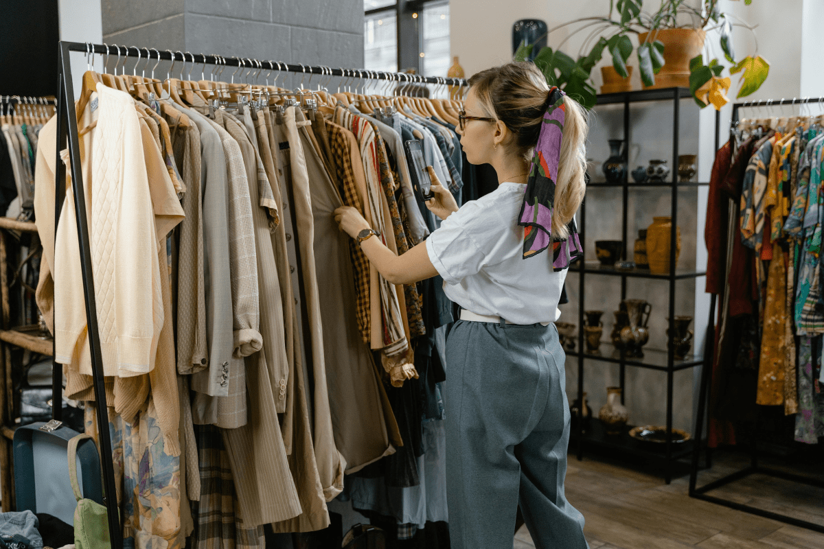 Sell and Resell - Side Hustle Apps A Woman is Standing Near a Rack of Clothes Selling Items Illustrating Side Hustle Apps