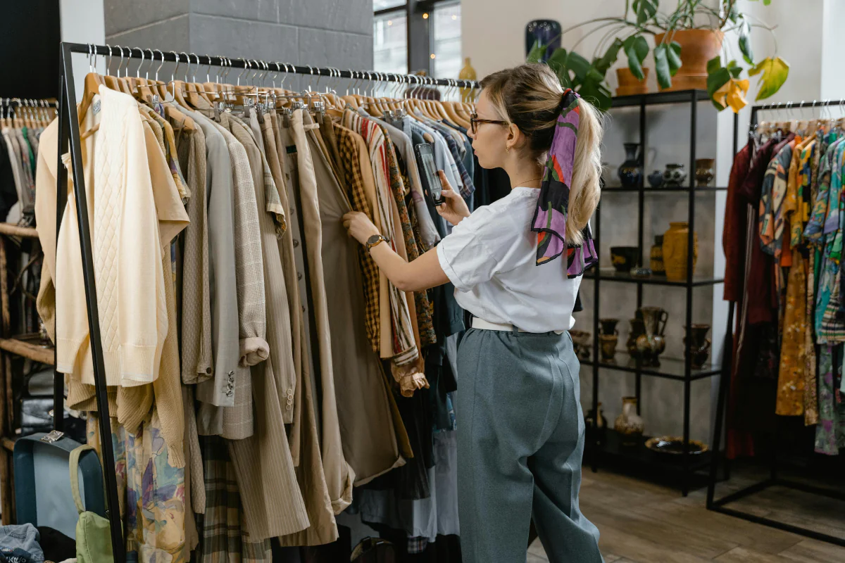 Sell and Resell - Side Hustle Apps A Woman is Standing Near a Rack of Clothes Selling Items Illustrating Side Hustle Apps