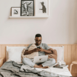 A Boy Using Their Phone with Their Laptop Lying in Their Lap as They Sit on Bed Illustrating Online Side Hustles from Home