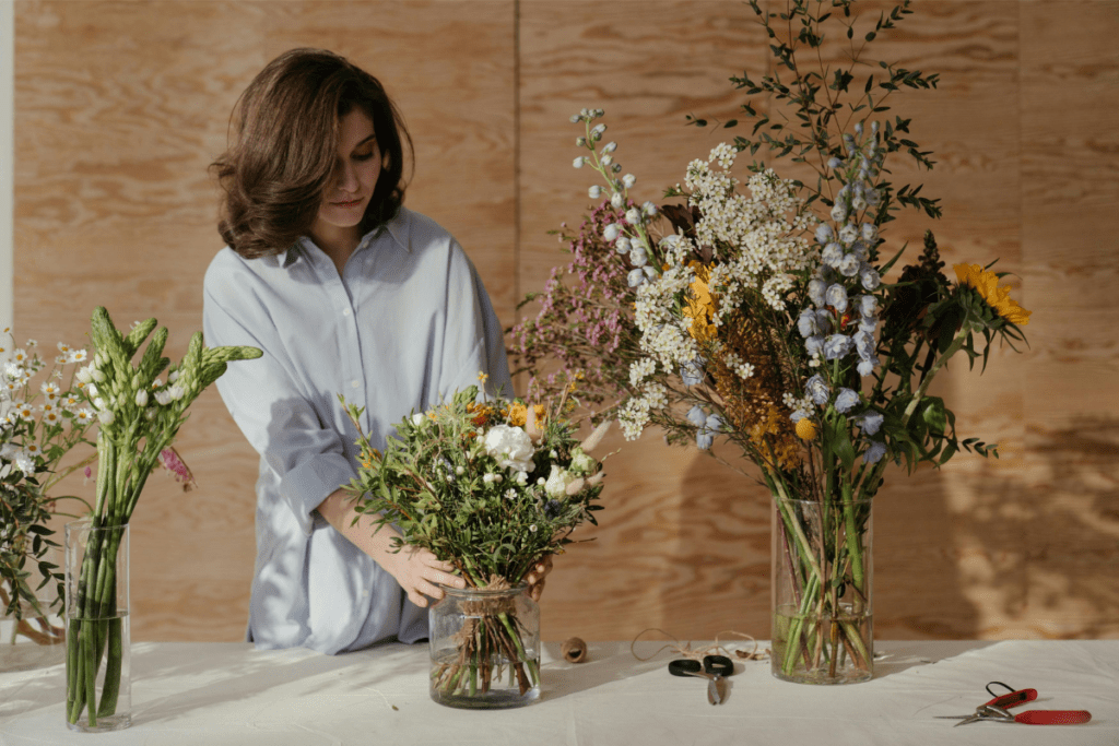 A Woman Can be Seen Arranging Flower in  A Vase, Three Vase are Placed on a Table Illustrating How to Start a Floral Business 