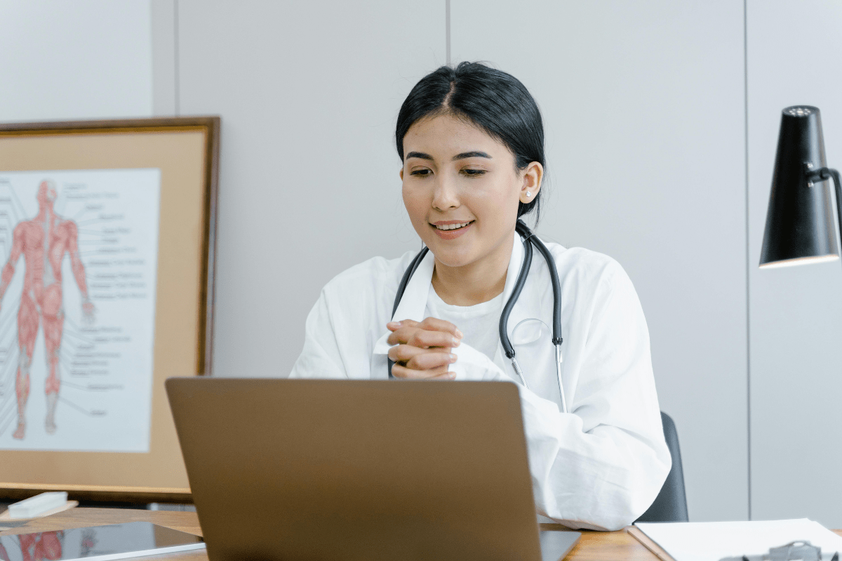 A Woman Wearing a Lab coat and a Stethoscope is Sitting in front of a Laptop Illustrating Pharmacist Side Hustle.