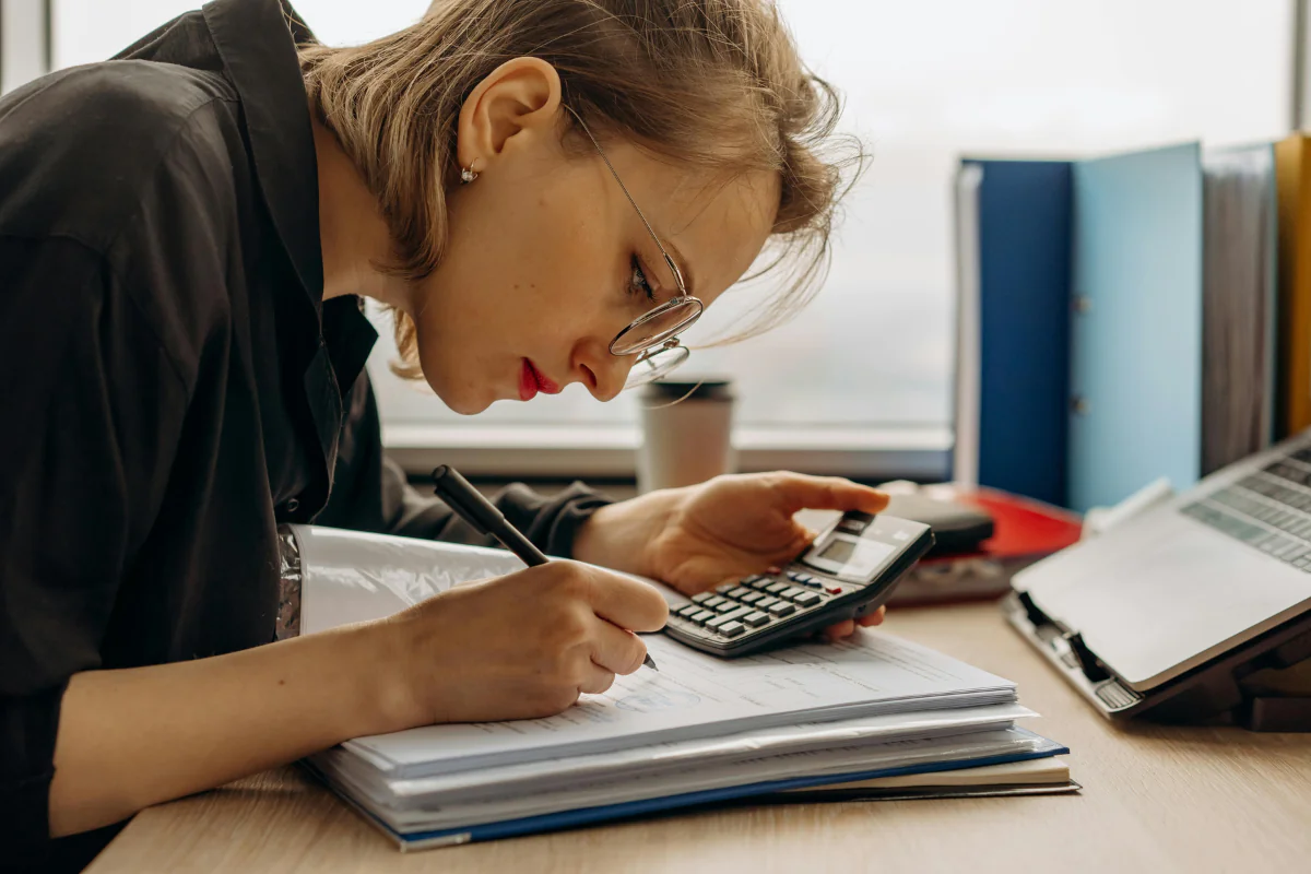 A Woman Writing Something in a Register While Holding a Calculator in Other Hand Illustrating Best Side Hustles for Accountants