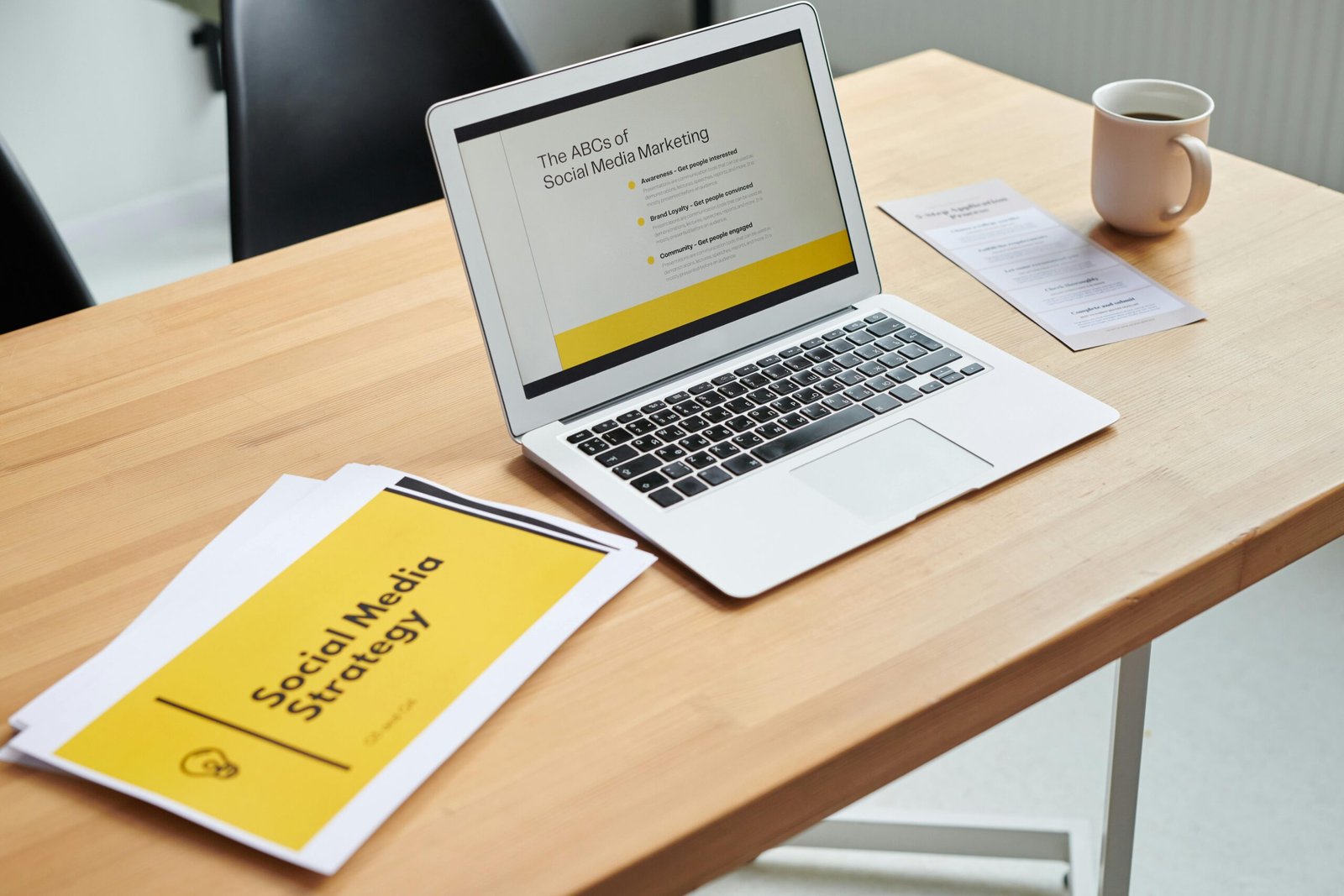 Promote Your Bakery - How to Start a Bakery A Laptop Is Placed on Table with "Social Media Strategy" Written on a Paper Besides It Illustrating How to Start a Bakery